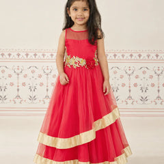 A young girl wearing a bright red sleeveless formal dress with gold floral embroidery at the waist and gold ruffled trim at the hem, standing against a light patterned wall.