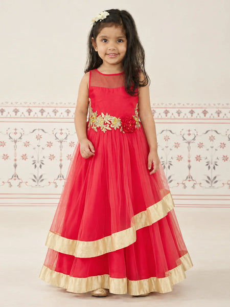 A young girl wearing a bright red sleeveless formal dress with gold floral embroidery at the waist and gold ruffled trim at the hem, standing against a light patterned wall.