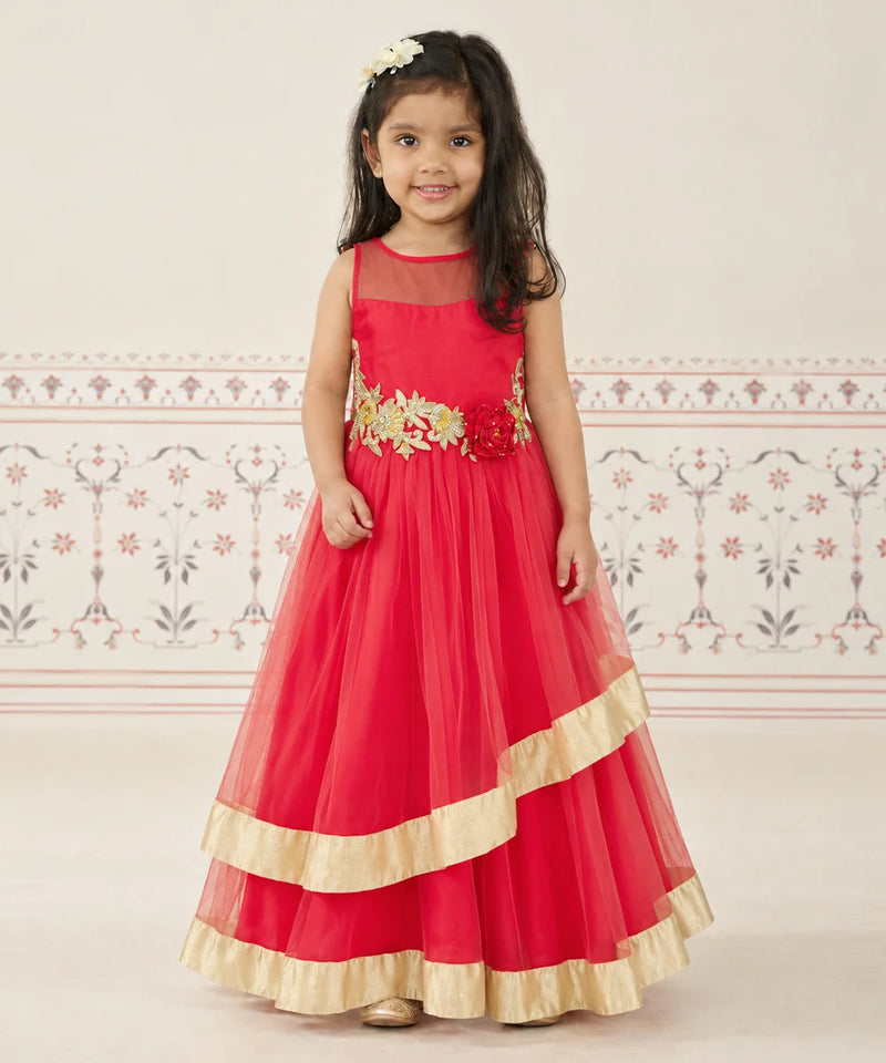 A young girl wearing a bright red sleeveless formal dress with gold floral embroidery at the waist and gold ruffled trim at the hem, standing against a light patterned wall.
