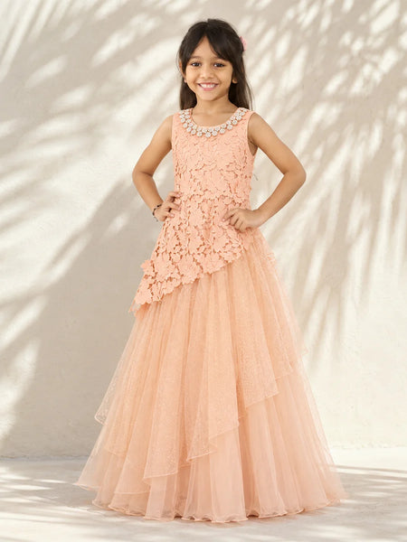 A young girl stands outdoors against a sunlit wall with leaf shadows, wearing a sleeveless peach formal dress with a beaded neckline, floral lace bodice, asymmetrical layered overlay, and a full tulle skirt.