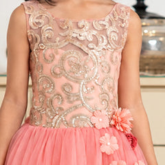 A close-up of a young girl wearing a sleeveless peach pink formal dress with a sheer embroidered bodice in metallic gold and a layered tulle skirt with pink fabric flowers.