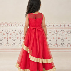 A young girl viewed from behind wearing a sleeveless red formal dress with a sheer back panel, a large back bow, and layered tulle with gold ruffle trim, standing against a light patterned wall.