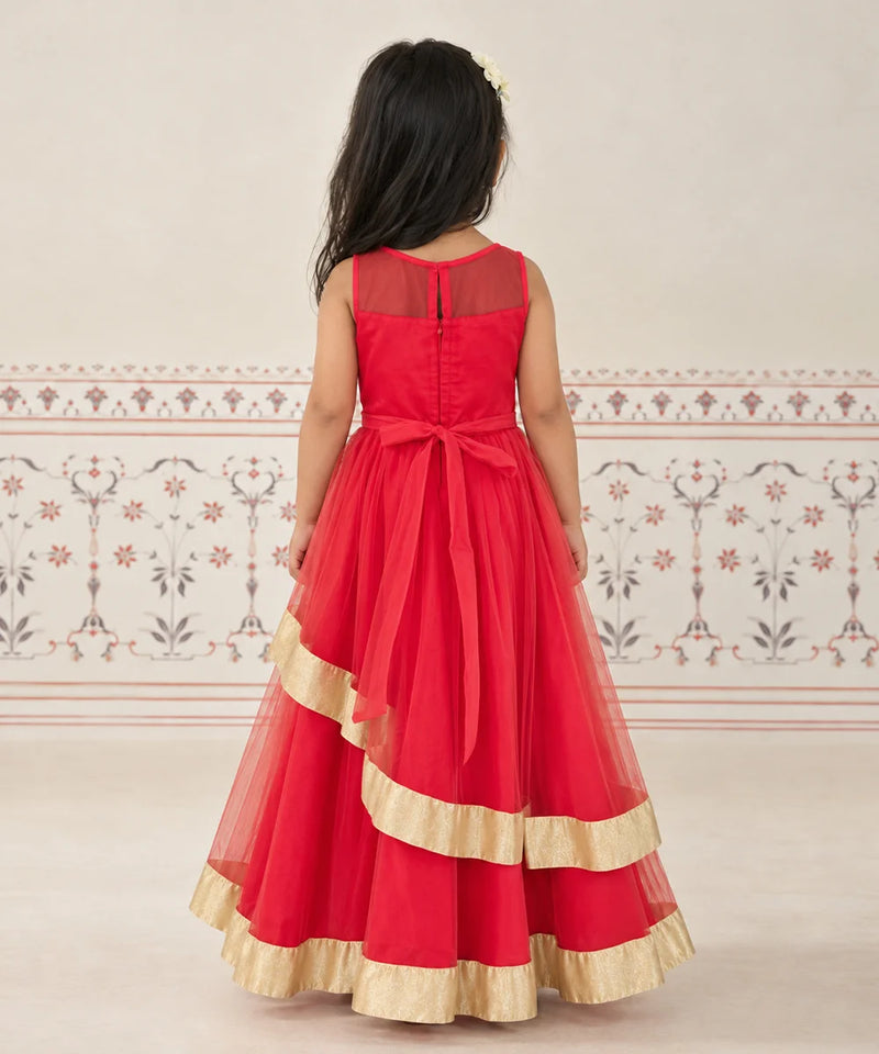 A young girl viewed from behind wearing a sleeveless red formal dress with a sheer back panel, a large back bow, and layered tulle with gold ruffle trim, standing against a light patterned wall.