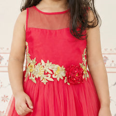 A young girl wearing a bright red sleeveless party dress with a sheer neckline, a tulle skirt, and a decorative gold floral waistband with a red flower accent.