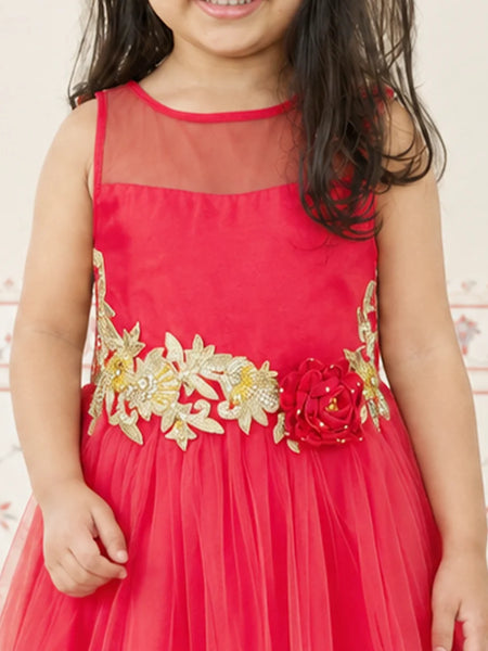 A young girl wearing a bright red sleeveless party dress with a sheer neckline, a tulle skirt, and a decorative gold floral waistband with a red flower accent.