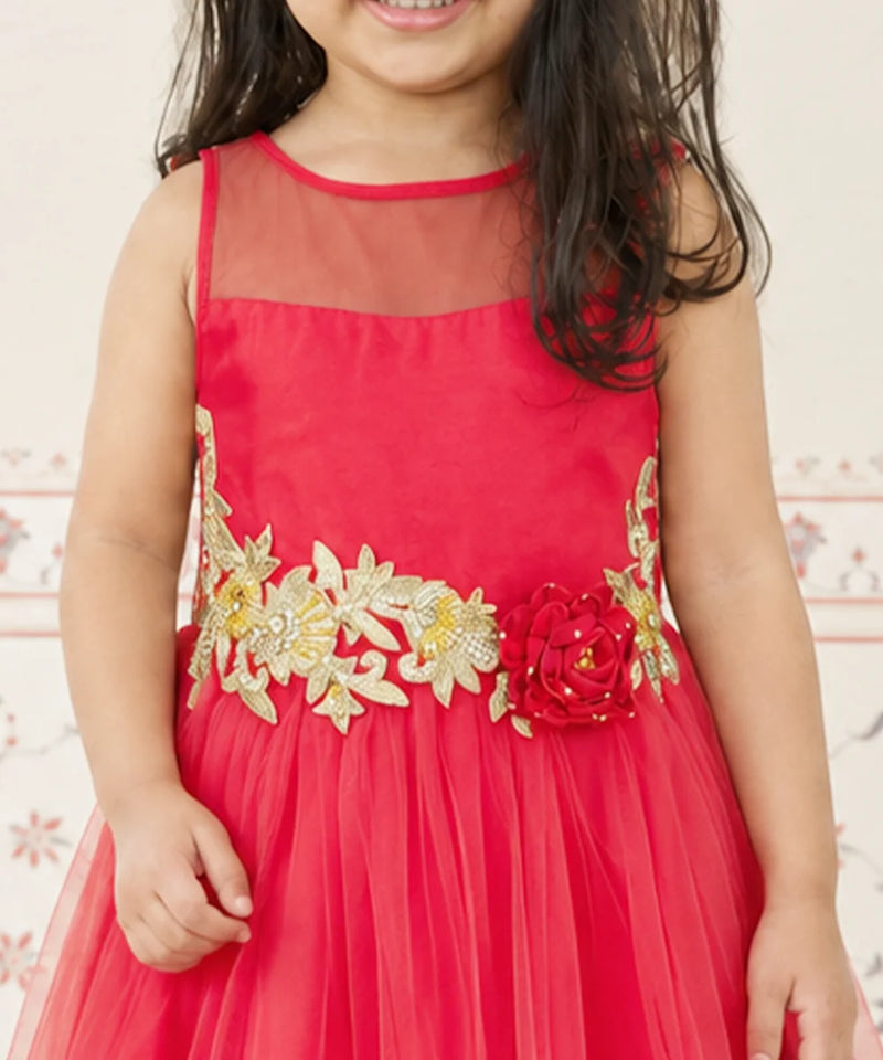 A young girl wearing a bright red sleeveless party dress with a sheer neckline, a tulle skirt, and a decorative gold floral waistband with a red flower accent.