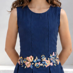 A sleeveless navy blue dress with vertical pleats and a colorful floral embroidered waistband, shown on a model against a light gray background.