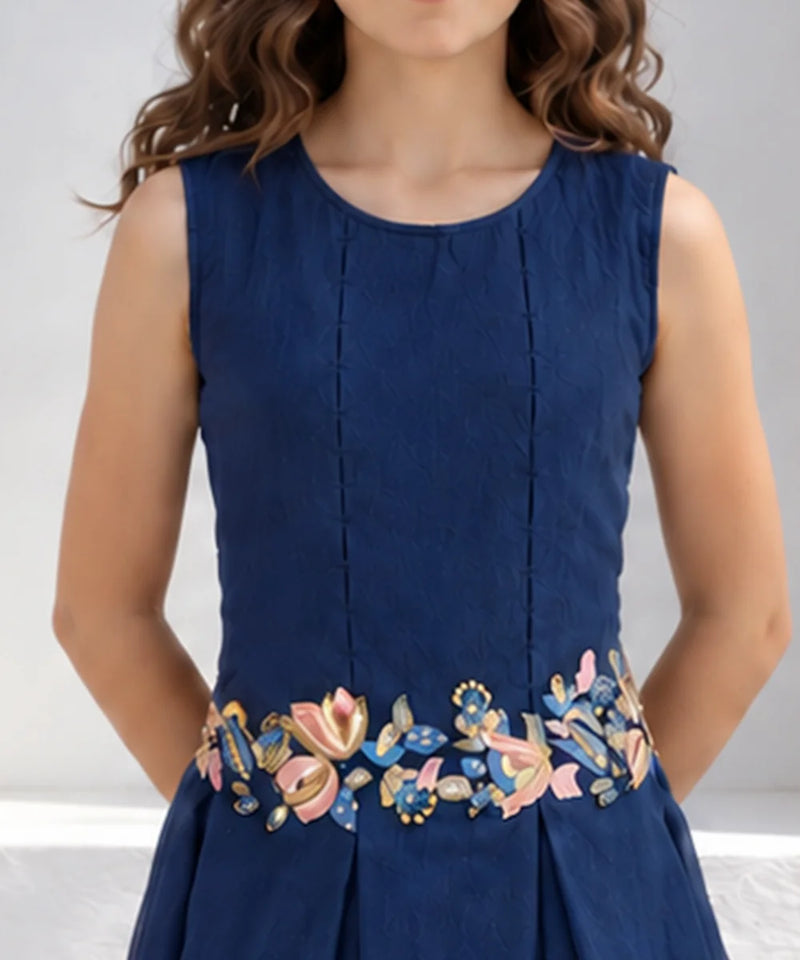 A sleeveless navy blue dress with vertical pleats and a colorful floral embroidered waistband, shown on a model against a light gray background.