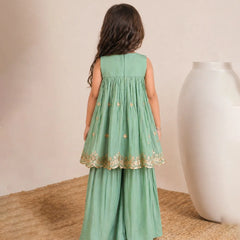 Young girl wearing a green dress with gold lace trim, standing on a textured surface with a neutral background.