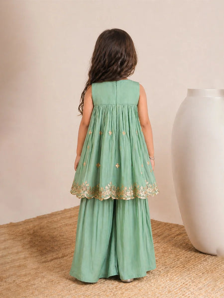 Young girl wearing a green dress with gold lace trim, standing on a textured surface with a neutral background.