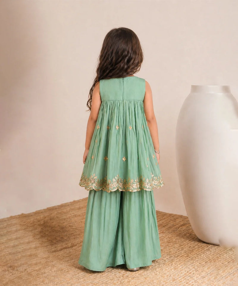 Young girl wearing a green dress with gold lace trim, standing on a textured surface with a neutral background.