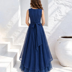 A woman stands with her back to the camera wearing a sleeveless navy blue A-line maxi dress with a matching fabric bow at the waist, set against a bright white stucco wall and white steps.