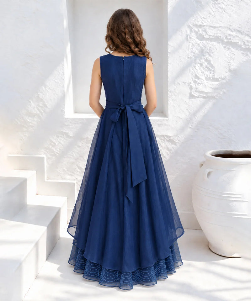 A woman stands with her back to the camera wearing a sleeveless navy blue A-line maxi dress with a matching fabric bow at the waist, set against a bright white stucco wall and white steps.