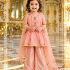 A young girl stands in a grand, ornate hall wearing a peach-pink traditional outfit with gold embroidery and a matching sheer dupatta.