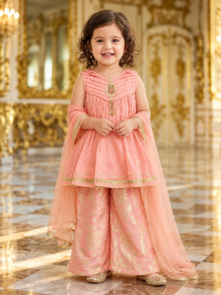 A young girl stands in a grand, ornate hall wearing a peach-pink traditional outfit with gold embroidery and a matching sheer dupatta.