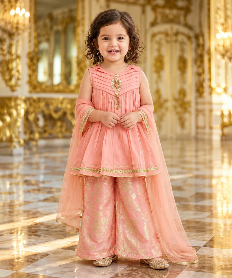 A young girl stands in a grand, ornate hall wearing a peach-pink traditional outfit with gold embroidery and a matching sheer dupatta.