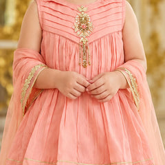 A child wearing a peach-pink traditional dress with gold embroidery and a matching sheer dupatta, shown from the shoulders to the knees against a softly blurred golden background.