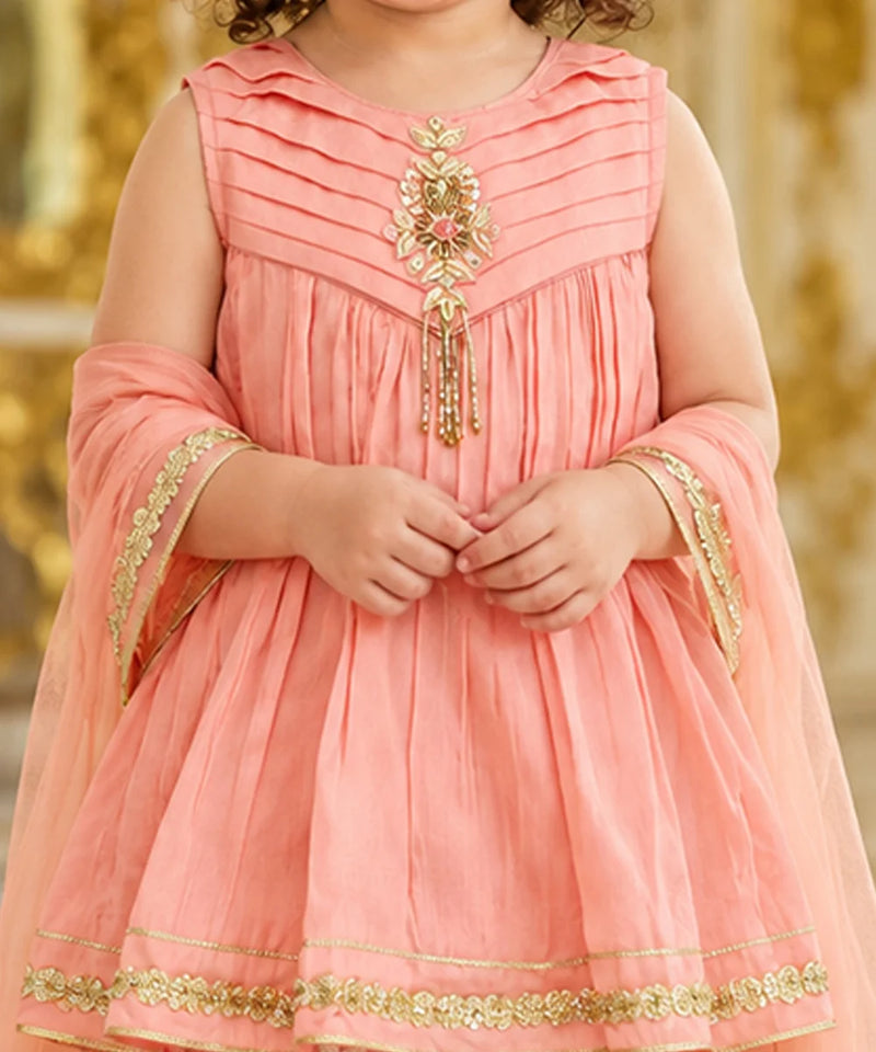 A child wearing a peach-pink traditional dress with gold embroidery and a matching sheer dupatta, shown from the shoulders to the knees against a softly blurred golden background.