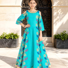 A young girl models a turquoise blue Anarkali gown with gold, pink, and blue floral patterns, standing in front of a beige stone building with an arched doorway.