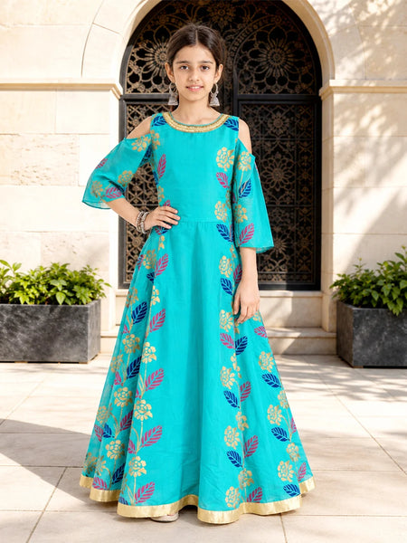 A young girl models a turquoise blue Anarkali gown with gold, pink, and blue floral patterns, standing in front of a beige stone building with an arched doorway.