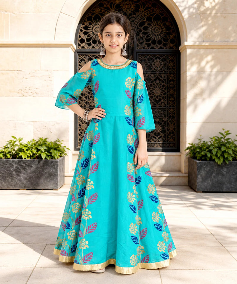 A young girl models a turquoise blue Anarkali gown with gold, pink, and blue floral patterns, standing in front of a beige stone building with an arched doorway.