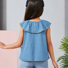 A child model shown from the back wearing a light blue denim sleeveless top with a ruffled collar and matching denim pants, set against a light gray wall with a pink block and a green plant.