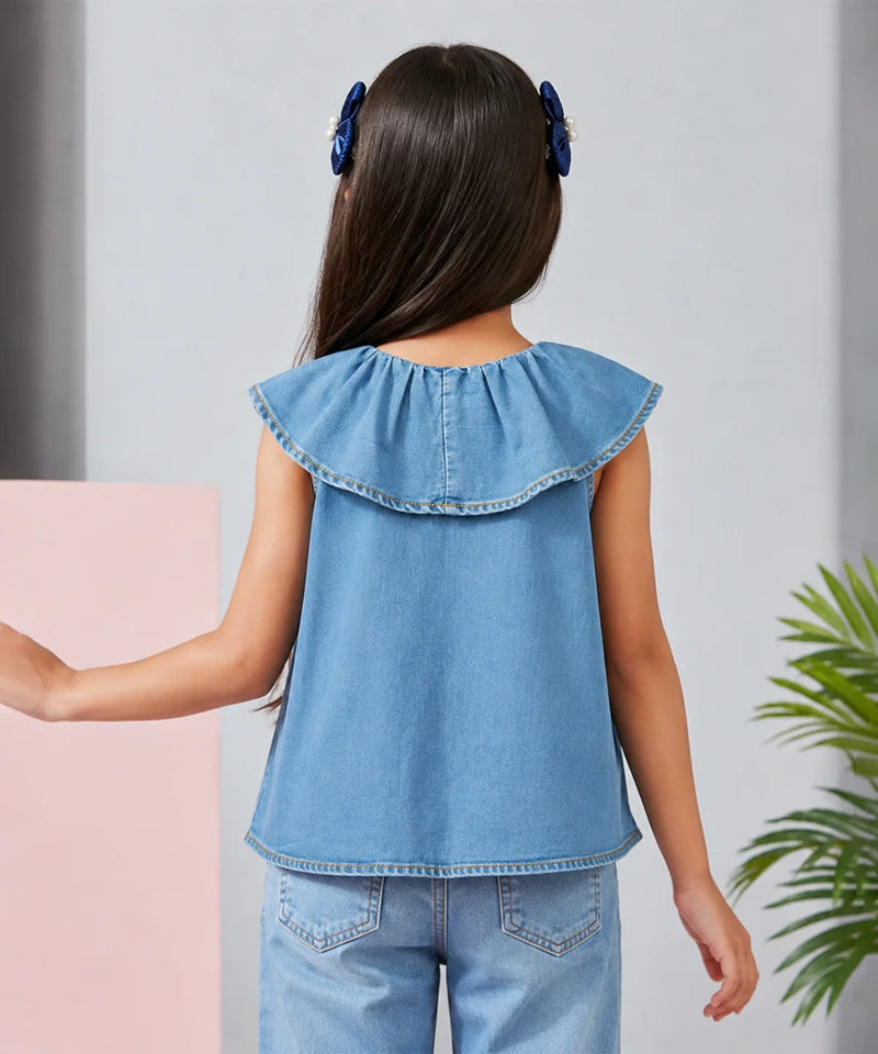 A child model shown from the back wearing a light blue denim sleeveless top with a ruffled collar and matching denim pants, set against a light gray wall with a pink block and a green plant.