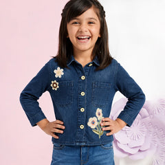 A young girl smiling and posing in a dark blue denim button-up shirt with embroidered flowers on the chest, paired with matching blue denim pants, set against a split pink and white background with decorative paper flowers.