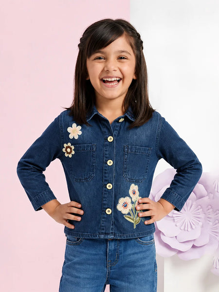 A young girl smiling and posing in a dark blue denim button-up shirt with embroidered flowers on the chest, paired with matching blue denim pants, set against a split pink and white background with decorative paper flowers.