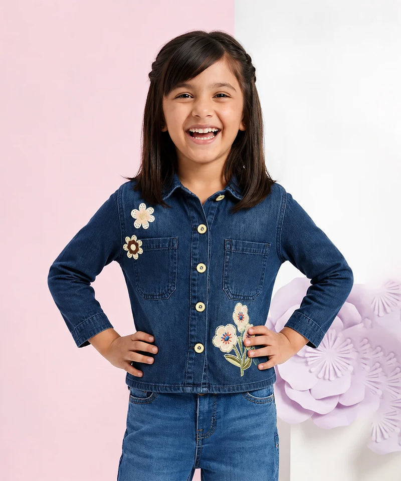 A young girl smiling and posing in a dark blue denim button-up shirt with embroidered flowers on the chest, paired with matching blue denim pants, set against a split pink and white background with decorative paper flowers.