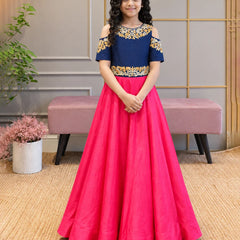 A young girl stands indoors wearing a formal off-the-shoulder gown with a navy blue bodice, gold floral embroidery, and a bright pink full skirt.