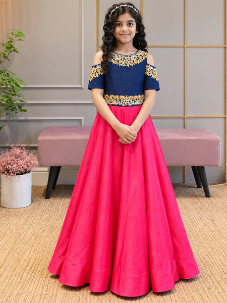 A young girl stands indoors wearing a formal off-the-shoulder gown with a navy blue bodice, gold floral embroidery, and a bright pink full skirt.