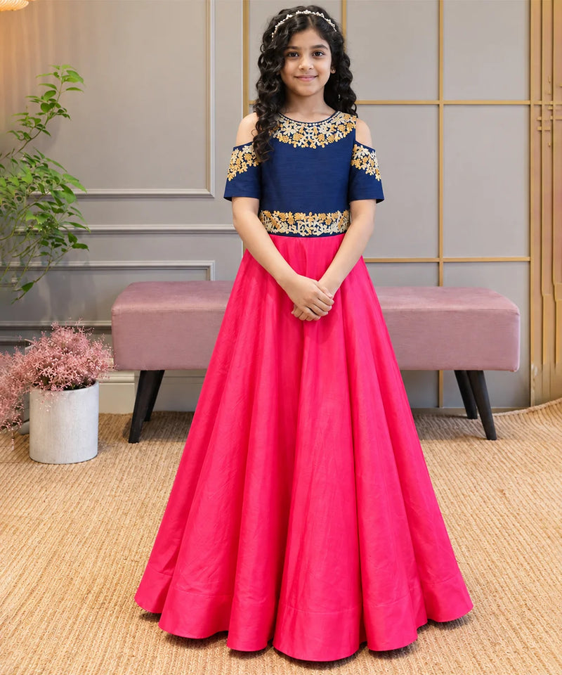 A young girl stands indoors wearing a formal off-the-shoulder gown with a navy blue bodice, gold floral embroidery, and a bright pink full skirt.