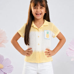 A young girl with long brown hair is smiling and posing in a two-tone yellow and white short-sleeve button-up shirt with a pineapple patch on the chest, paired with white shorts, set against a light gray background with decorative pink flowers.
