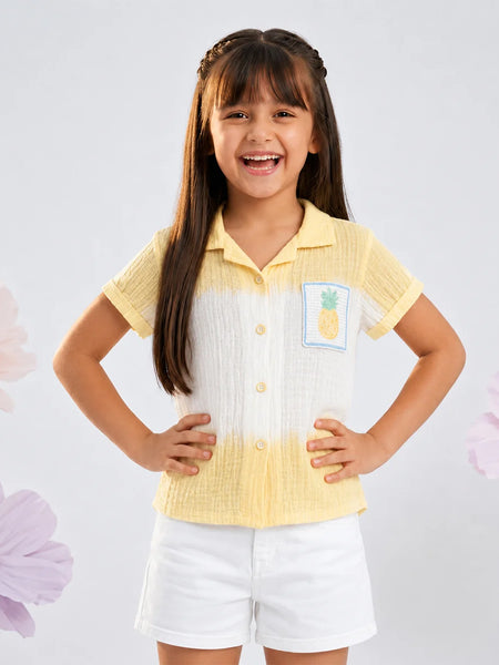 A young girl with long brown hair is smiling and posing in a two-tone yellow and white short-sleeve button-up shirt with a pineapple patch on the chest, paired with white shorts, set against a light gray background with decorative pink flowers.