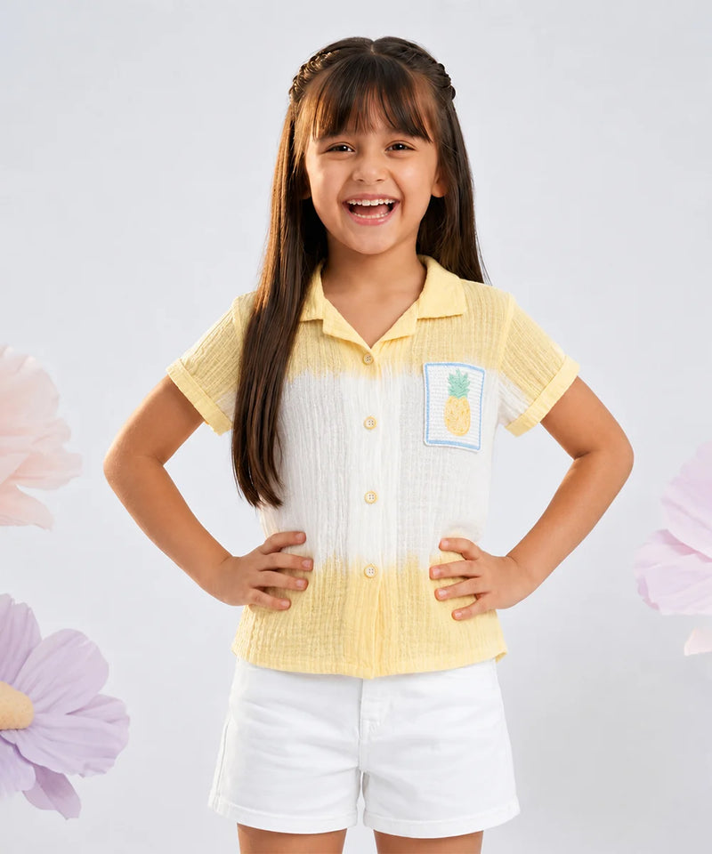 A young girl with long brown hair is smiling and posing in a two-tone yellow and white short-sleeve button-up shirt with a pineapple patch on the chest, paired with white shorts, set against a light gray background with decorative pink flowers.