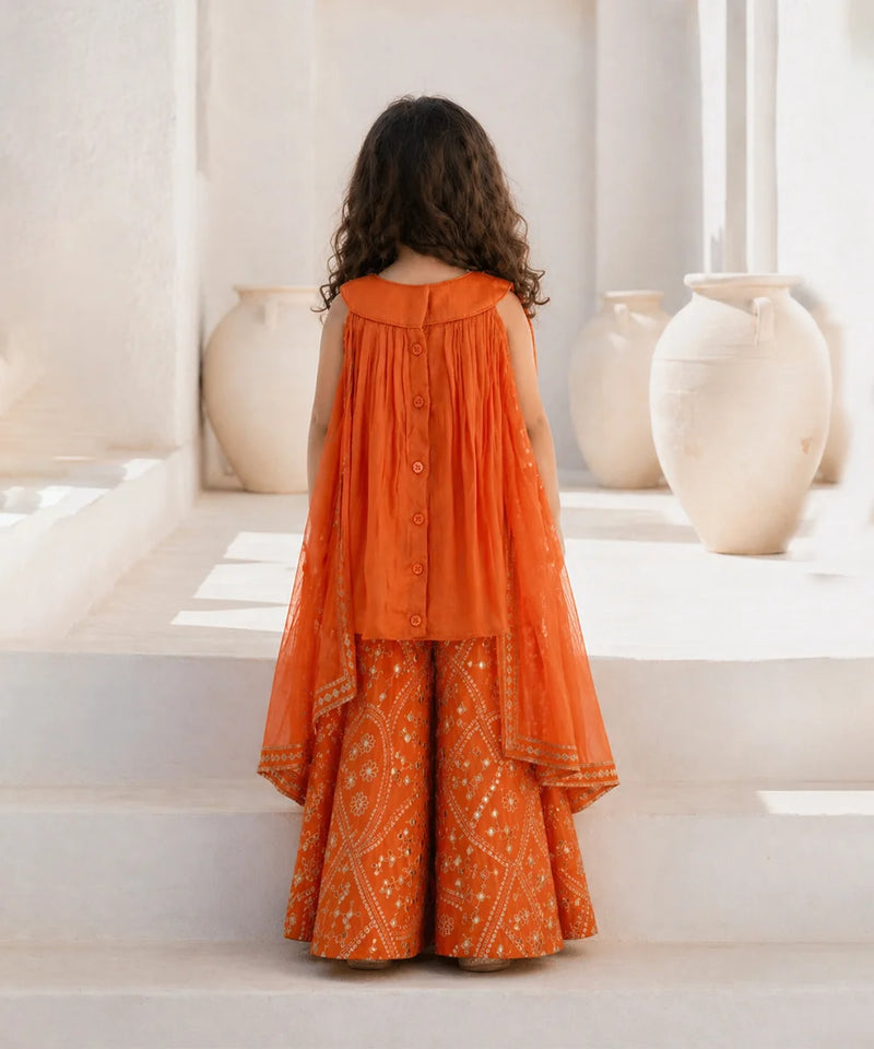 Person wearing an orange traditional outfit standing on a white staircase with decorative vases in the background.