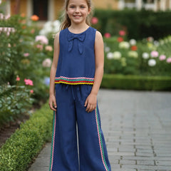 A young girl stands on a stone path in a garden wearing a navy blue sleeveless jumpsuit with a bow at the neckline, a colorful striped hem detail, and matching wide-leg pants with side stripes.