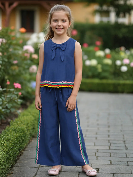 A young girl stands on a stone path in a garden wearing a navy blue sleeveless jumpsuit with a bow at the neckline, a colorful striped hem detail, and matching wide-leg pants with side stripes.