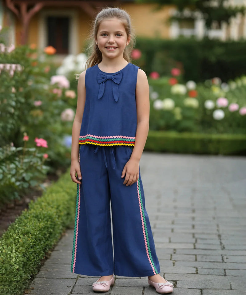 A young girl stands on a stone path in a garden wearing a navy blue sleeveless jumpsuit with a bow at the neckline, a colorful striped hem detail, and matching wide-leg pants with side stripes.