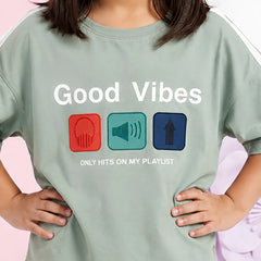 A child wearing a sage green graphic t-shirt with the text 'Good Vibes ONLY HITS ON MY PLAYLIST' and three colored icons representing headphones, a speaker, and an arrow up.
