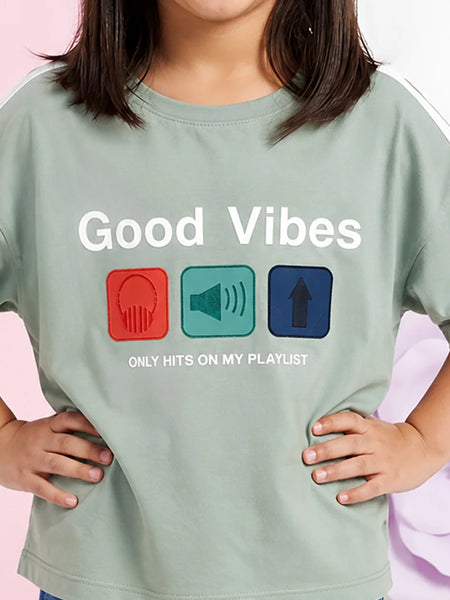 A child wearing a sage green graphic t-shirt with the text 'Good Vibes ONLY HITS ON MY PLAYLIST' and three colored icons representing headphones, a speaker, and an arrow up.