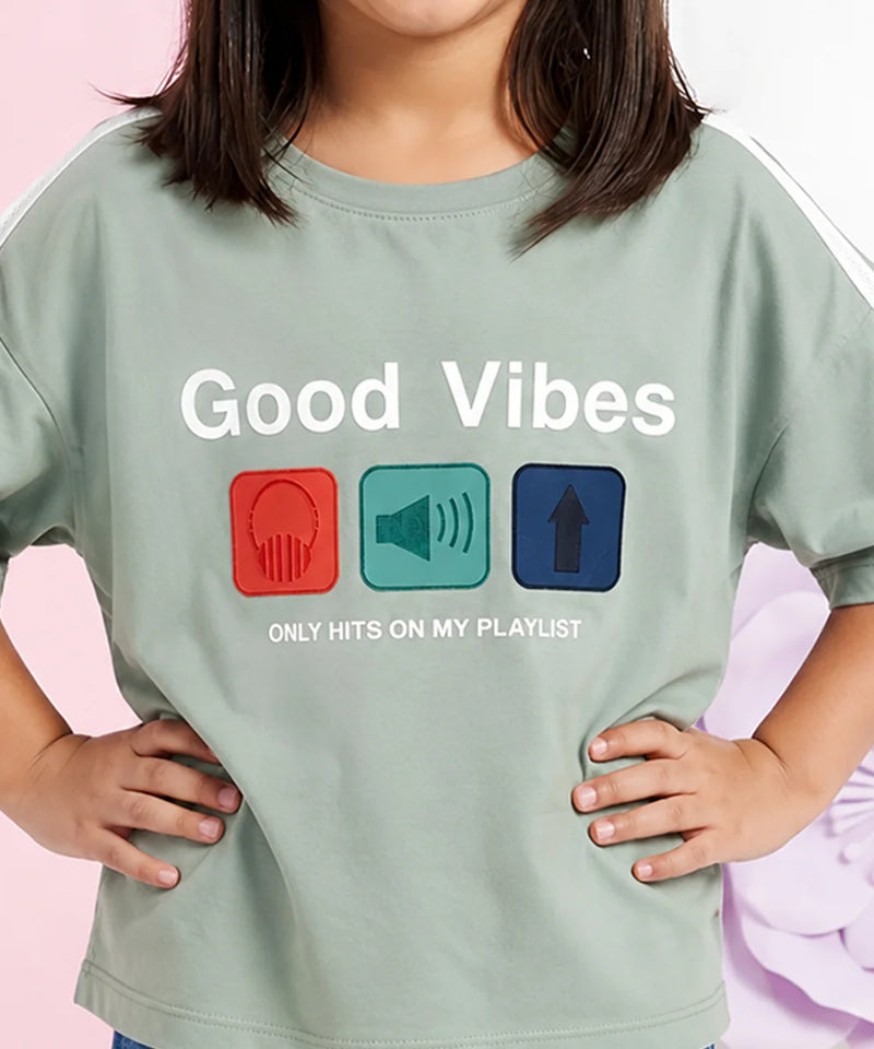 A child wearing a sage green graphic t-shirt with the text 'Good Vibes ONLY HITS ON MY PLAYLIST' and three colored icons representing headphones, a speaker, and an arrow up.