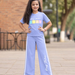 A young girl stands outdoors wearing a light blue matching tracksuit set consisting of a short-sleeve t-shirt and wide-leg pants. The t-shirt features 'Plajy Stadium' text and a colorful block graphic, and the pants have white side stripes and 'Plajy Stadium' text down the leg.