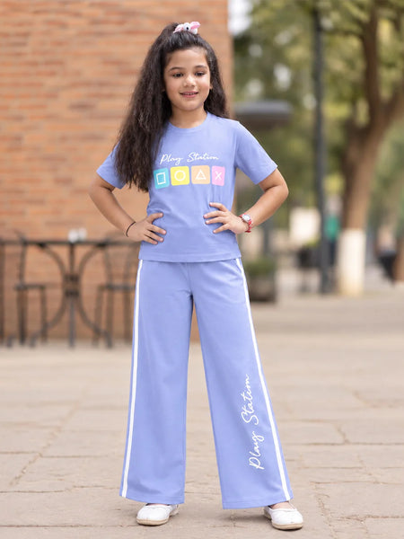A young girl stands outdoors wearing a light blue matching tracksuit set consisting of a short-sleeve t-shirt and wide-leg pants. The t-shirt features 'Plajy Stadium' text and a colorful block graphic, and the pants have white side stripes and 'Plajy Stadium' text down the leg.
