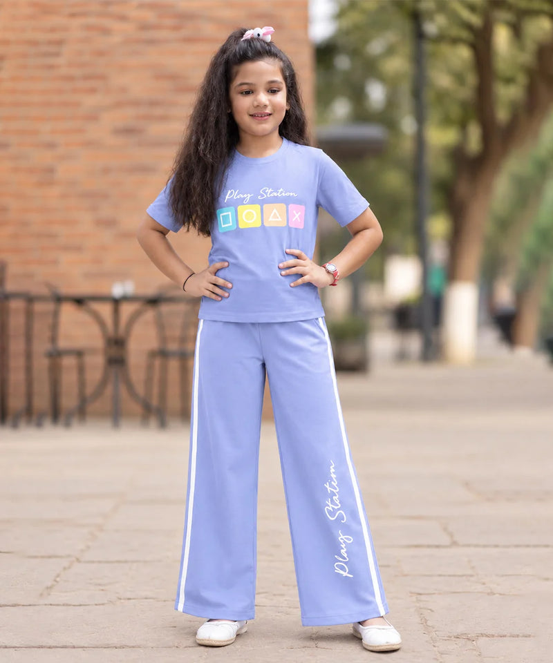 A young girl stands outdoors wearing a light blue matching tracksuit set consisting of a short-sleeve t-shirt and wide-leg pants. The t-shirt features 'Plajy Stadium' text and a colorful block graphic, and the pants have white side stripes and 'Plajy Stadium' text down the leg.