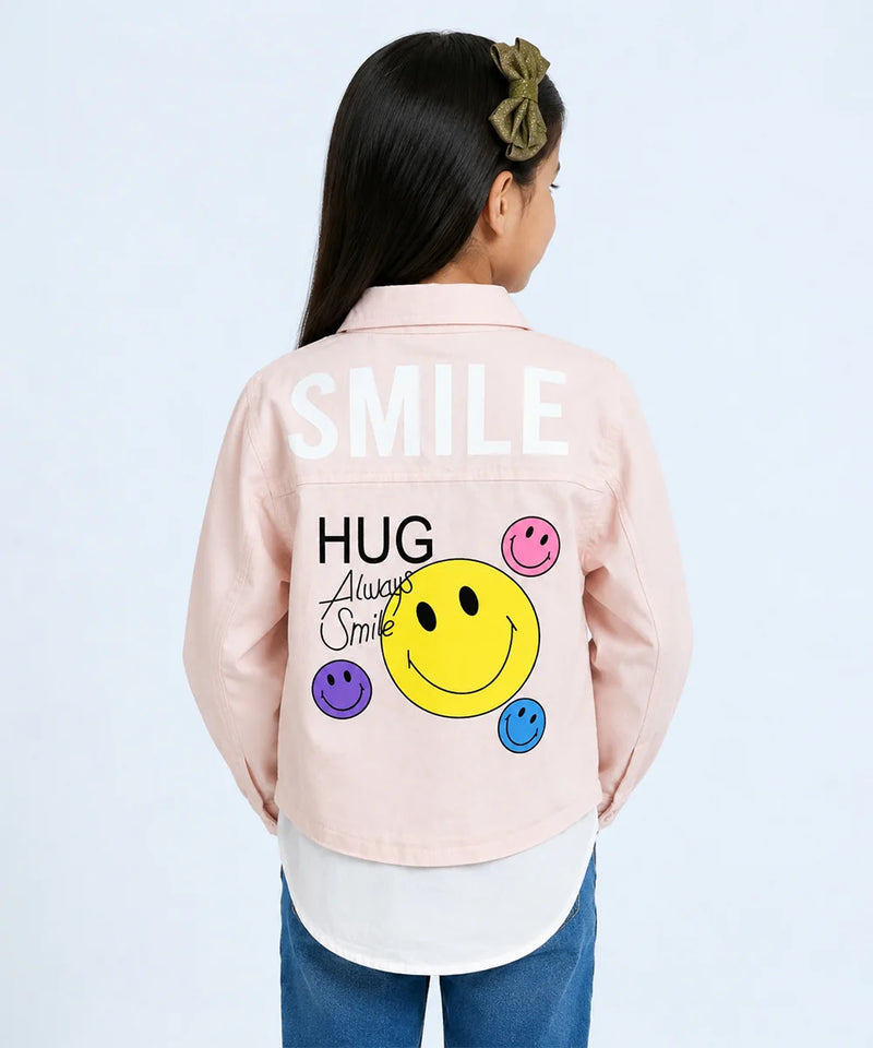 A child model shown from the back wearing a light pink denim-style jacket with a large graphic on the back. The graphic features the word 'SMILE' in white, 'HUG Always Smile' in black script and block lettering, a large yellow smiley face, and smaller pink, purple, and blue smiley faces.