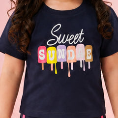A child wearing a navy blue t-shirt with a colorful graphic that reads 'Sweet SUNDAE' in dripping popsicle shapes, set against a solid pink background.