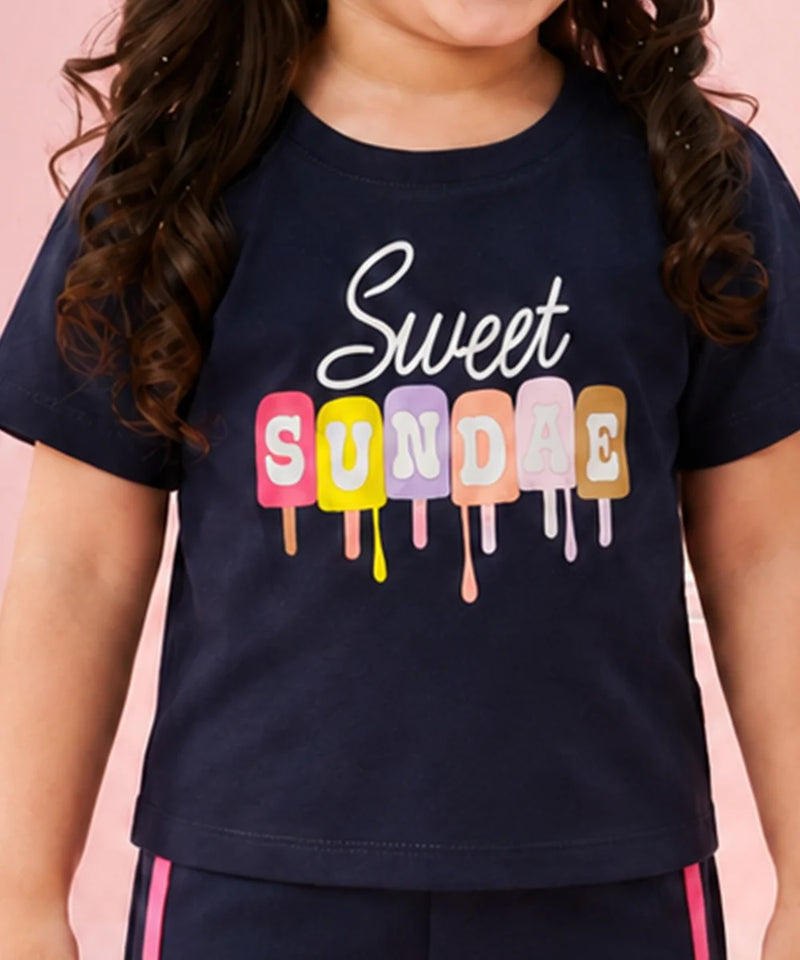 A child wearing a navy blue t-shirt with a colorful graphic that reads 'Sweet SUNDAE' in dripping popsicle shapes, set against a solid pink background.