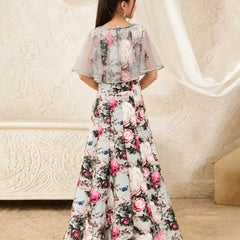 A woman viewed from behind wearing a long floral printed gown with sheer flutter sleeves and a flared skirt, standing indoors against a cream textured wall and draped fabric.